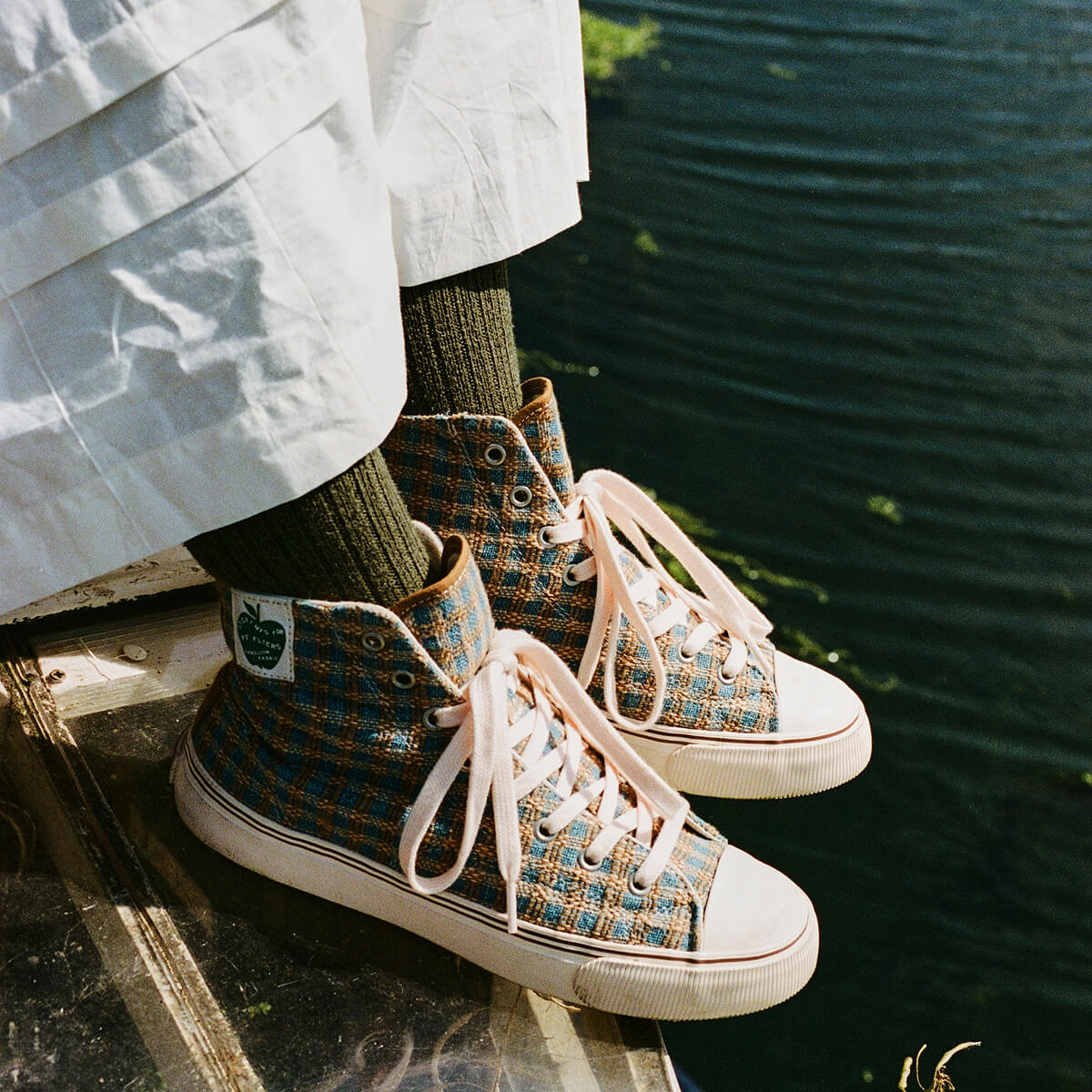 Person sitting beside dark water wearing indigo and brown plaid high-top sneakers.
