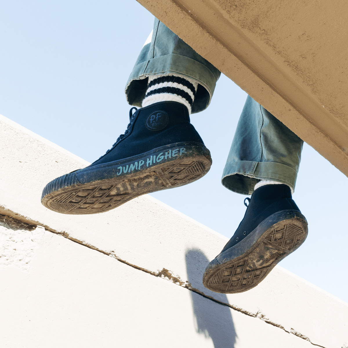 Person wearing black sneakers with 'Jump Higher' text, sitting on a ledge with a clear sky background.