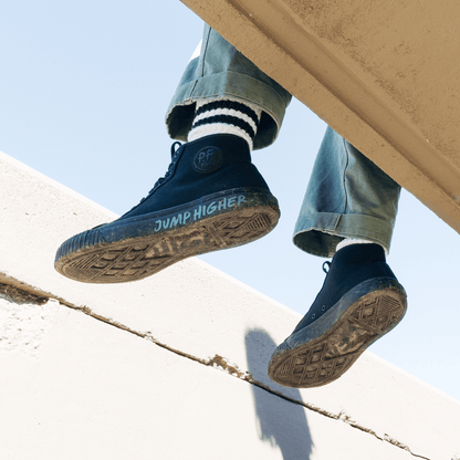 Person wearing black sneakers with 'Jump Higher' text, sitting on a ledge with a clear sky background.