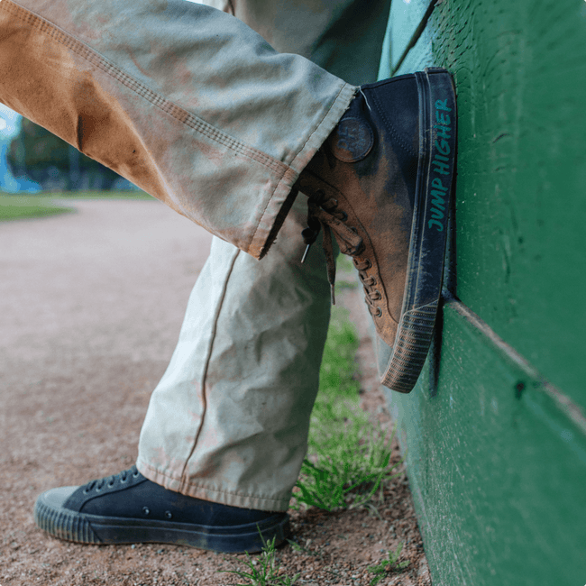 Person wearing black high-top shoes with 'Jump Higher' text, leaning against a green wall outdoors.