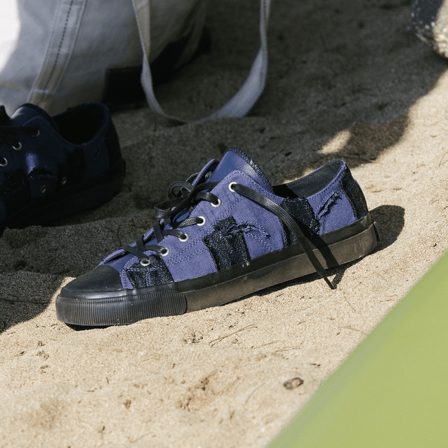 Navy low-top sneakers with black soles on a sandy surface.