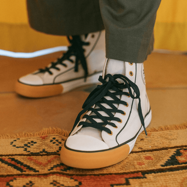 White retro high-top sneakers with black laces and gum soles, shown on a person standing on a patterned rug.