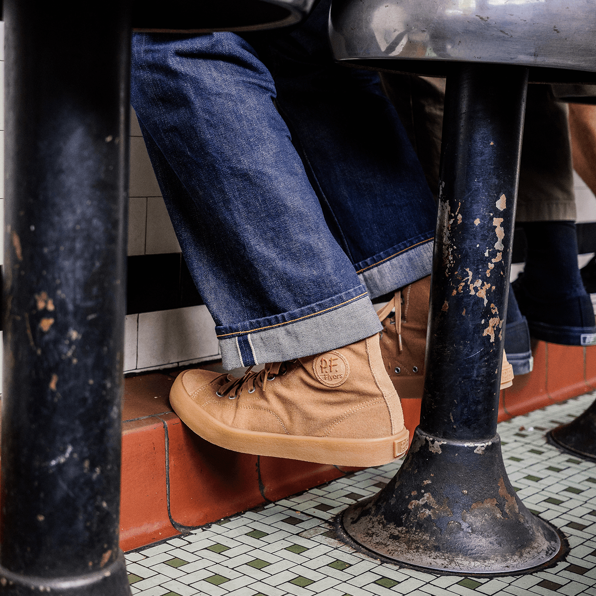 Person seated on a metal stool wearing tan high-top sneakers and blue jeans.