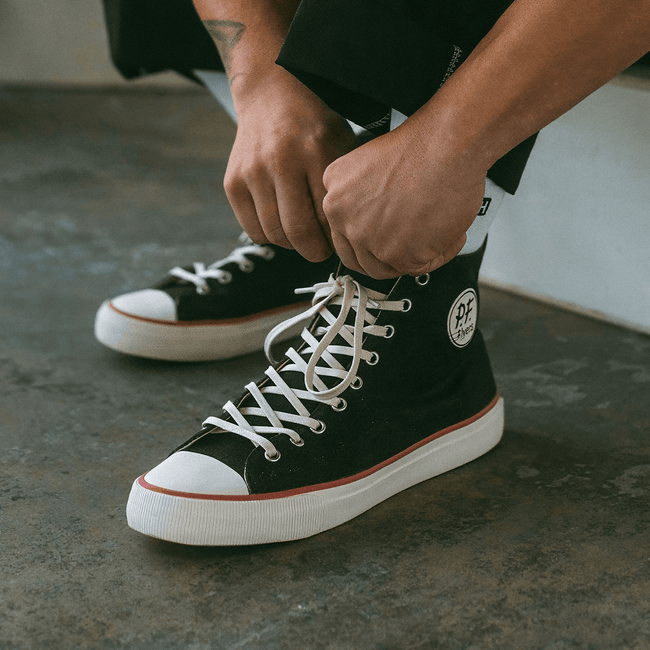 Person tying black high-top sneakers with white laces on a concrete floor.
