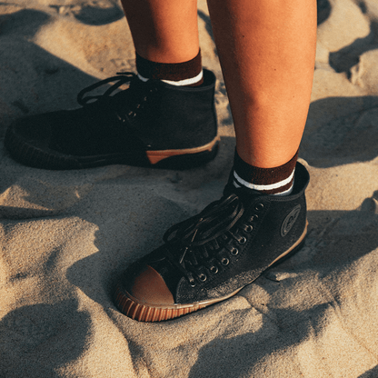 Person wearing black high-top sneakers with brown soles on sand.