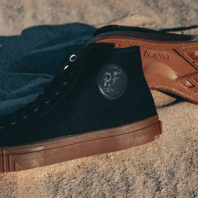 Black high-top sneakers with brown soles on a sandy surface.