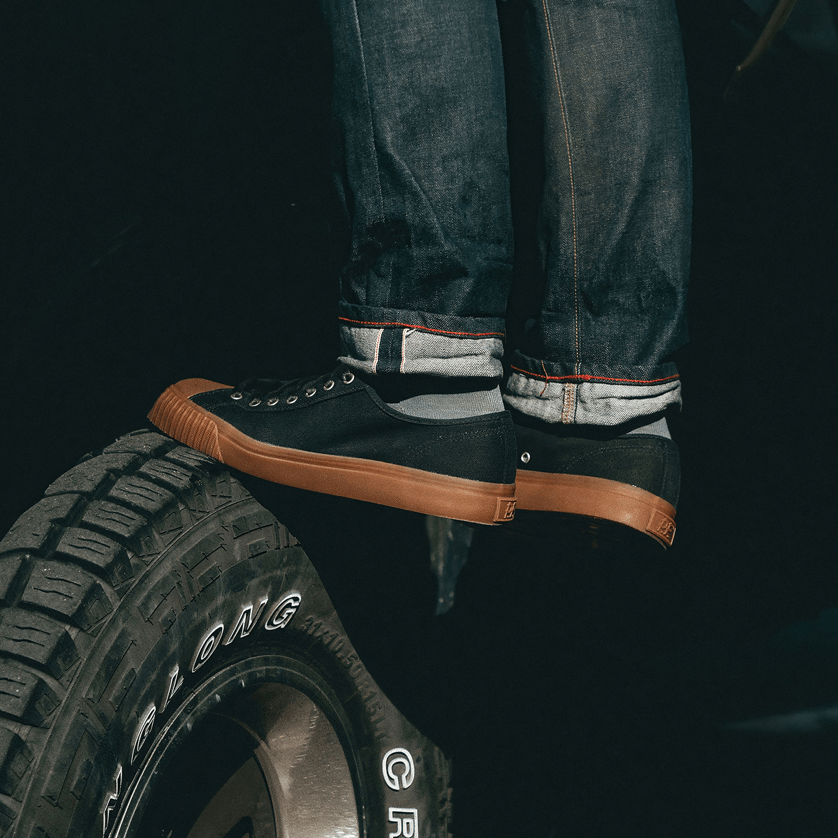 Person standing on a tire wearing black low-top sneakers with brown soles.