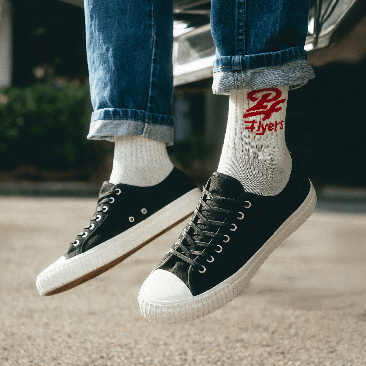 Black low-top sneakers with white soles worn with rolled-up jeans and white socks featuring a P.F. Flyers retro logo.