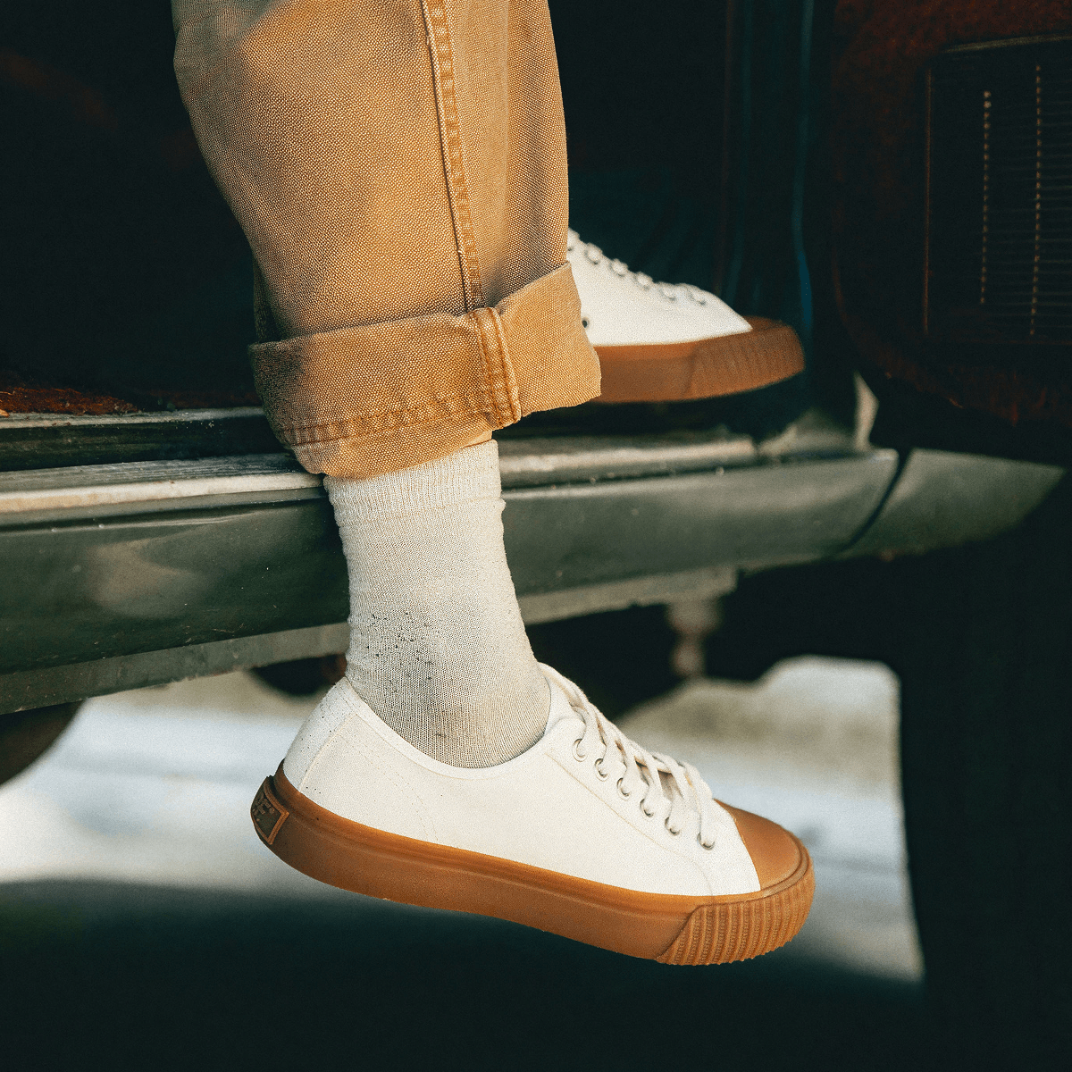 White low-top sneakers with brown soles worn by a person sitting inside of a vehicle.