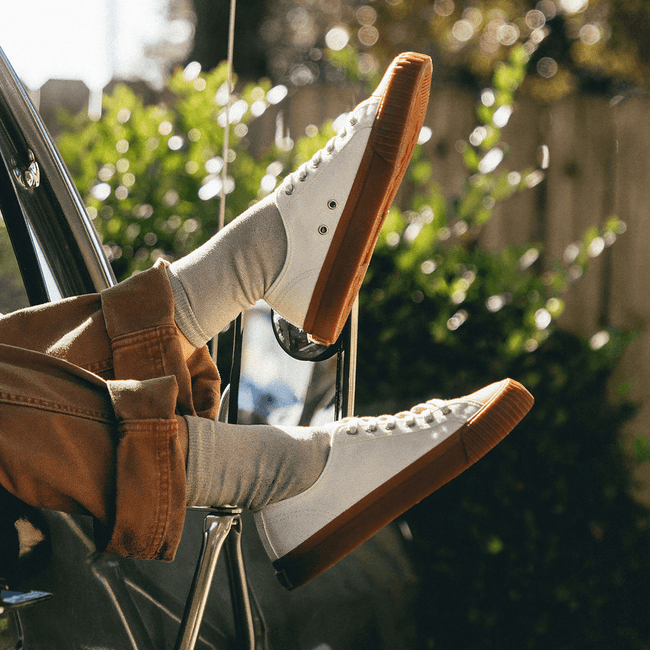 Person sitting in a car with their feet out the window, wearing white low-top sneakers with brown soles.