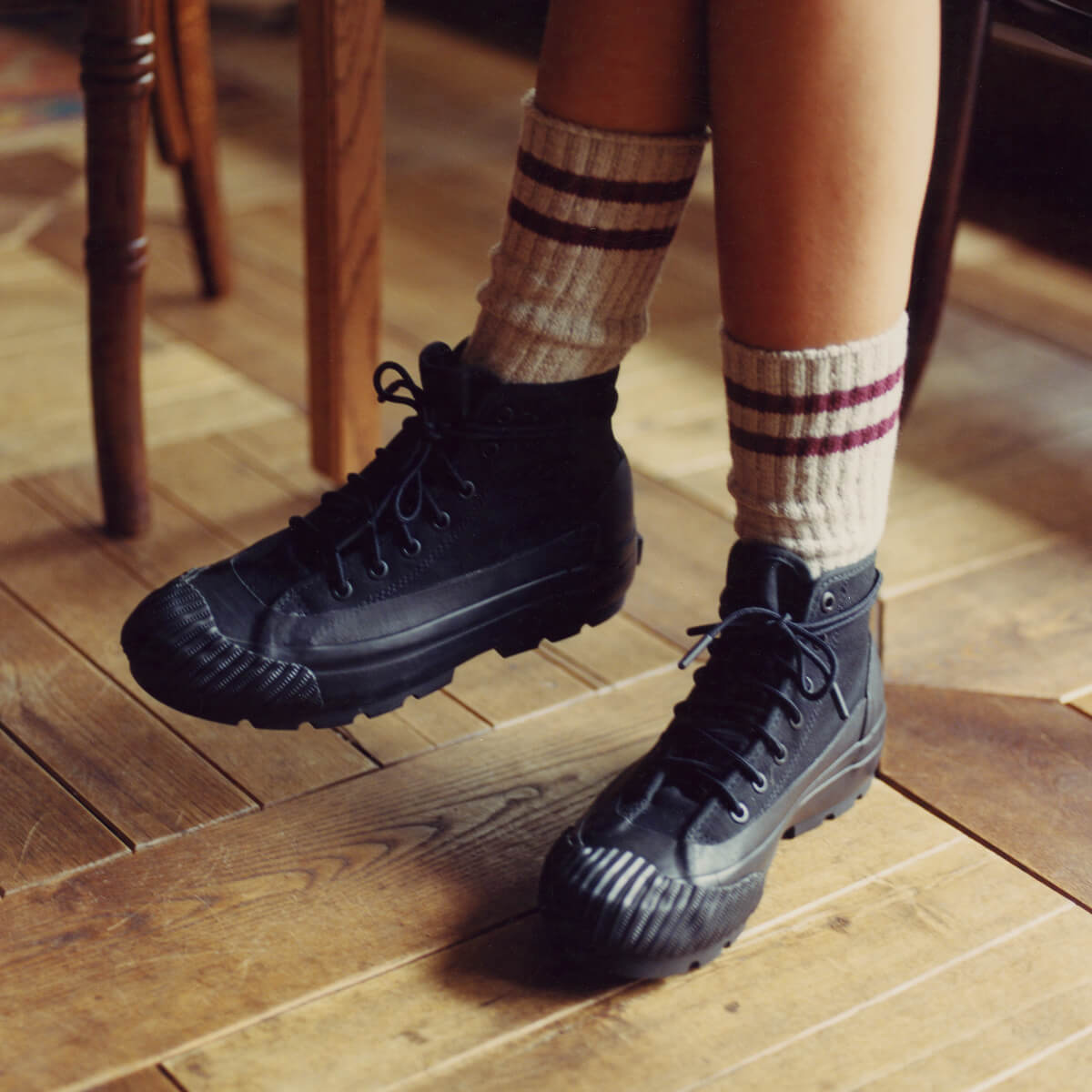 A person wearing black canvas boots with lugged rubber outsoles on a wooden floor.
