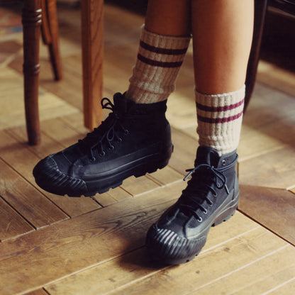 A person wearing black canvas boots with lugged rubber outsoles on a wooden floor.