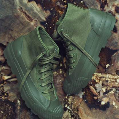 A pair of olive green boots with lugged rubber outsoles on a forest floor.