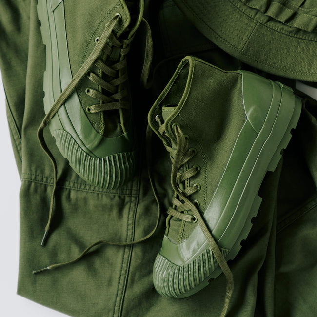 A pair of olive green boots with lugged rubber outsoles, displayed on top of an olive green pants against a light gray background.