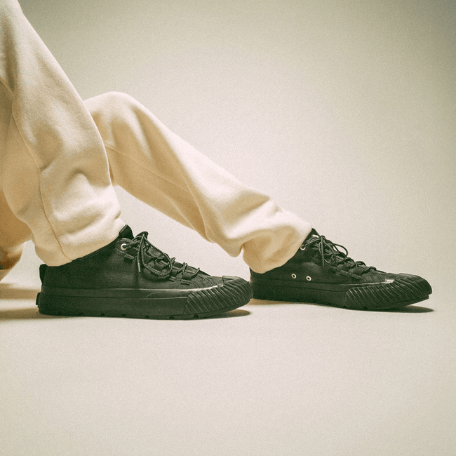 Black low-top sneakers worn with beige sweatpants on a neutral background.