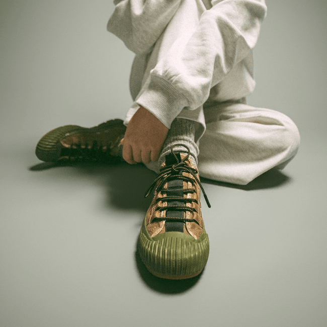 Person sitting on a gray floor with one knee raised, wearing a khaki-and-green sneaker.