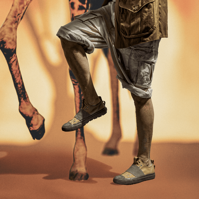 Person wearing khaki-and-green slip-on shoes and shorts standing beside a giraffe against a warm-toned background.