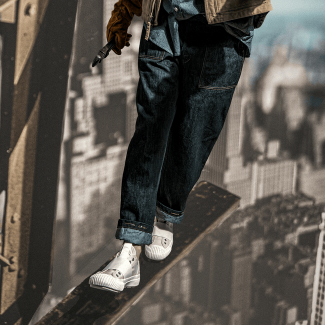 Person wearing blue jeans and white slip-on shoes standing on a construction beam with a cityscape in the background.