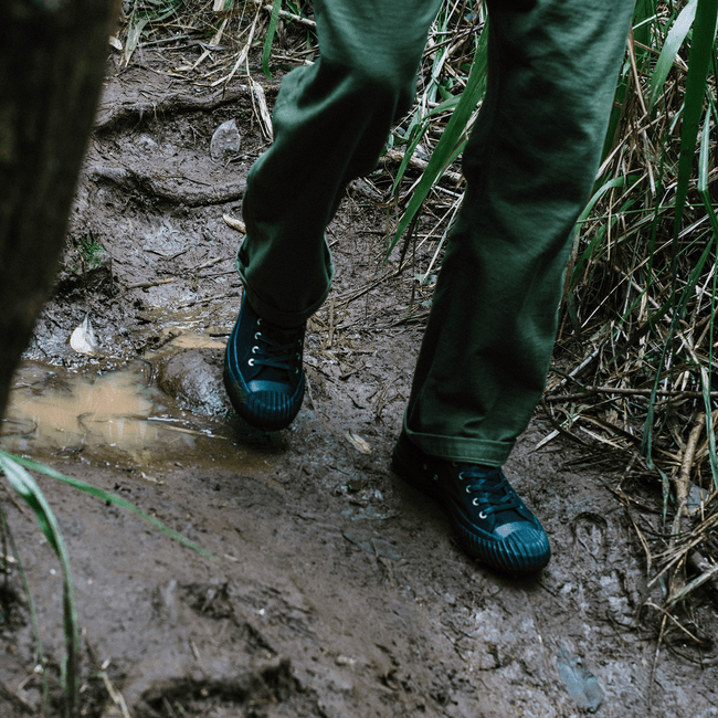 Person wearing green pants and black high-top sneakers walking along a muddy path.