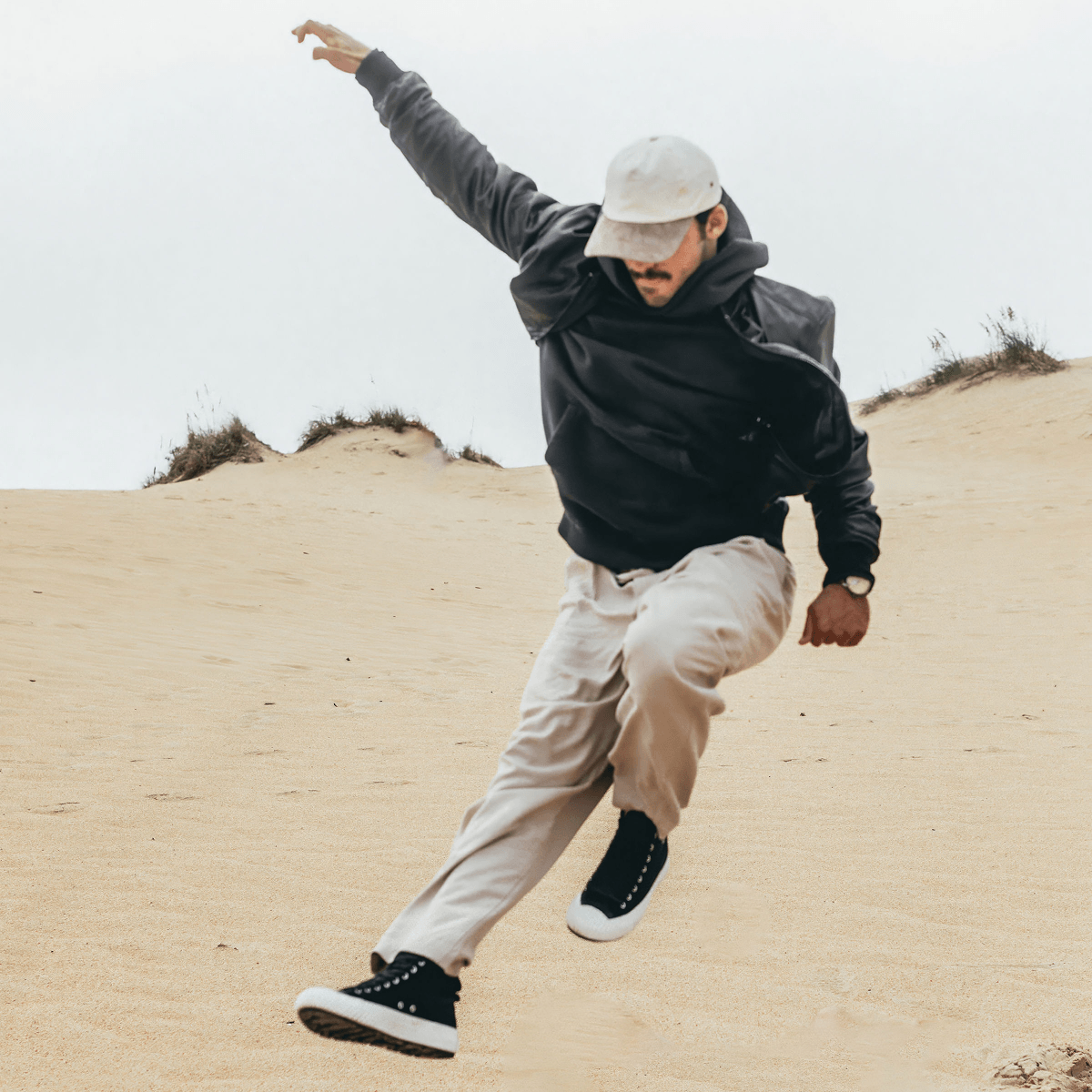 Person wearing casual clothing and black-and-white high-top sneakers leaping on sand dunes.