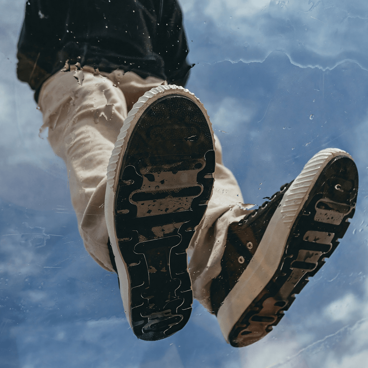 Person wearing black and white sneakers with beige pants against a blue sky.