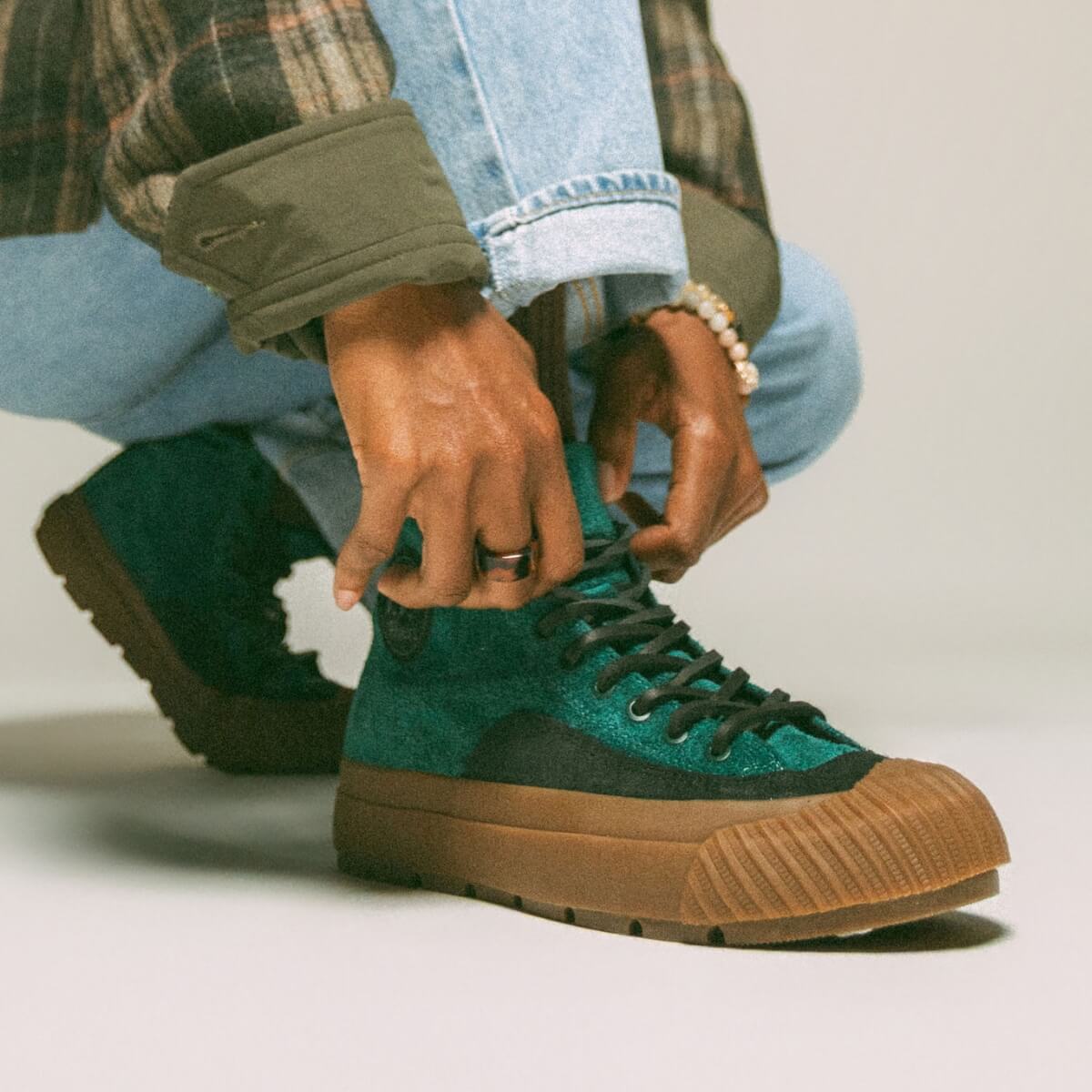Person tying a green and black sneaker with plaid shirt and jeans.