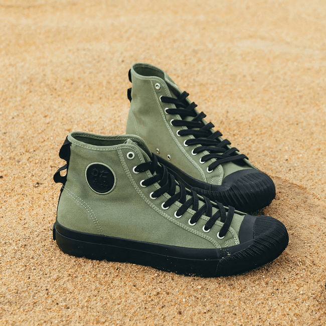 Green high-top sneakers with black soles on a sandy surface.