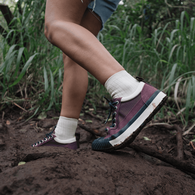Person wearing burgundy low-top sneakers on a muddy trail with greenery in the background.