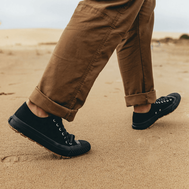 Person wearing black low-top sneakers and brown pants walking on a sand dune.