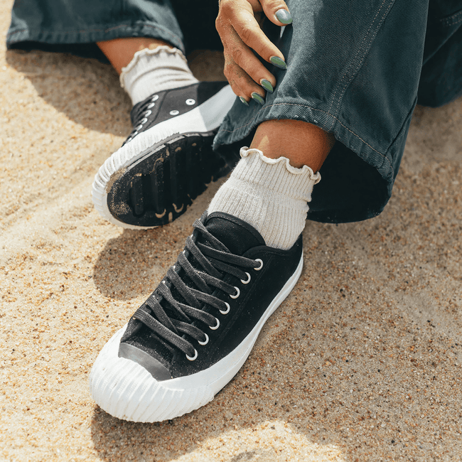 Black sneakers with white soles worn on a sandy surface.