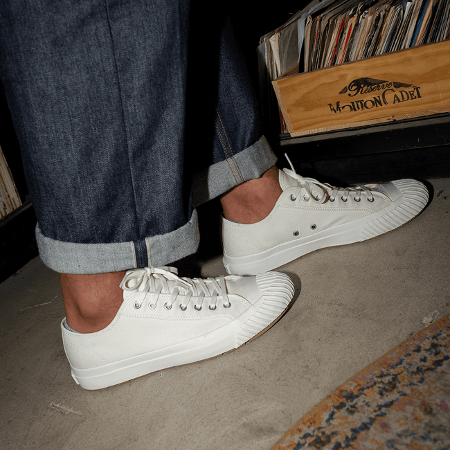 Person wearing white low-top sneakers with rolled-up jeans standing on a concrete floor.