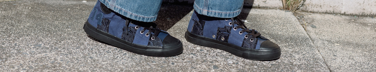 Navy high-top sneakers with black patches worn with jeans on a concrete surface.