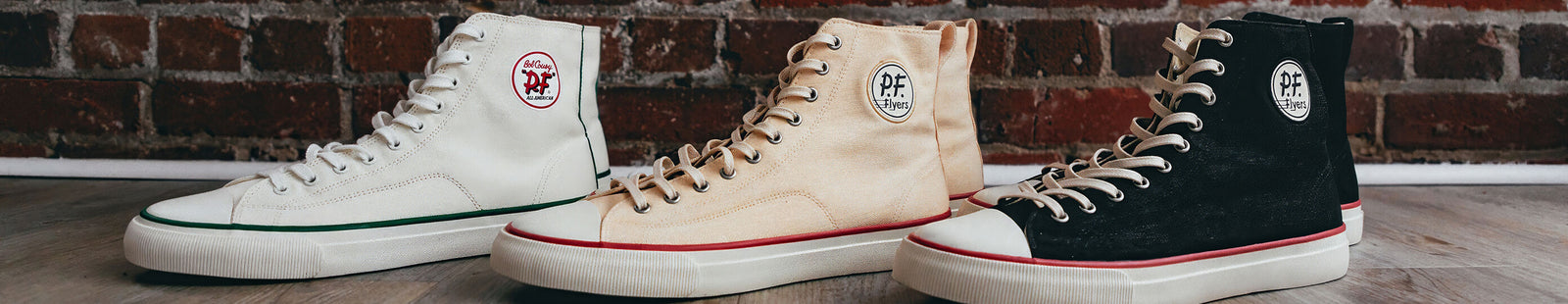 Three pairs of high-top sneakers in white, beige, and black against a brick wall background.
