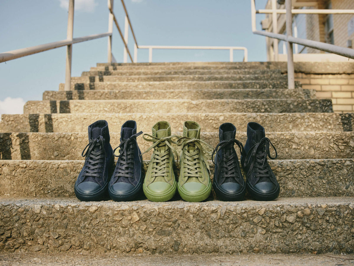 Three pairs of navy, olive green, and black high-top sneakers arranged on a concrete step under a blue sky.