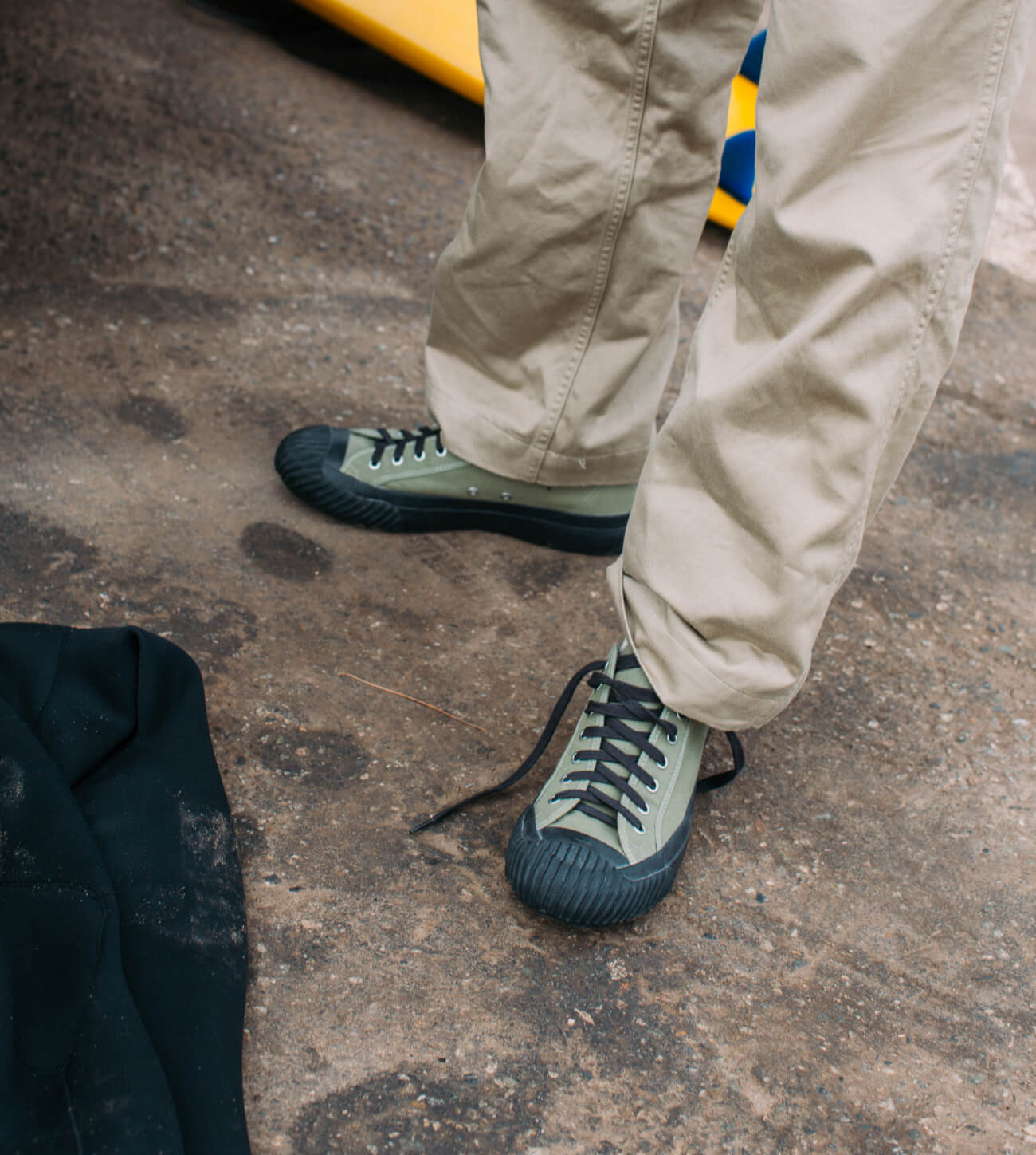 A person wearing beige pants and light green low-top canvas sneakers with dark laces and dark rubber toe caps, standing on a textured concrete ground.