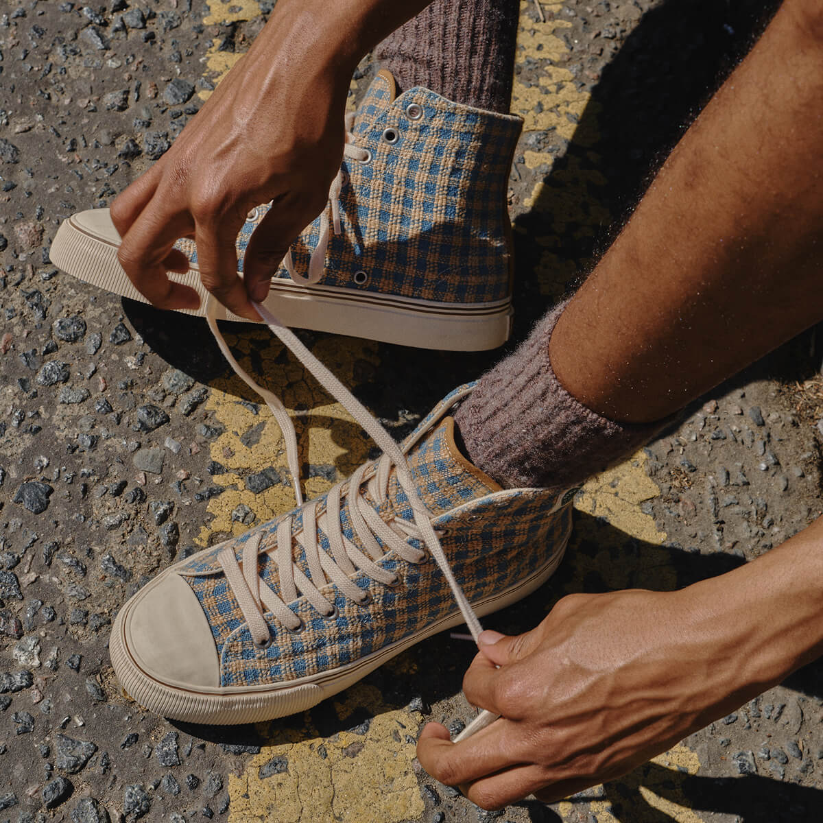 Person tying the laces of an indigo and brown plaid high-top sneaker, set against an asphalt background.