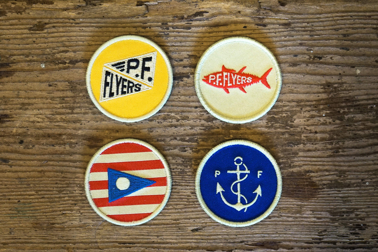 Four round patches with different designs on a wooden surface. P.F. Flyers x Slightly Choppy Collab